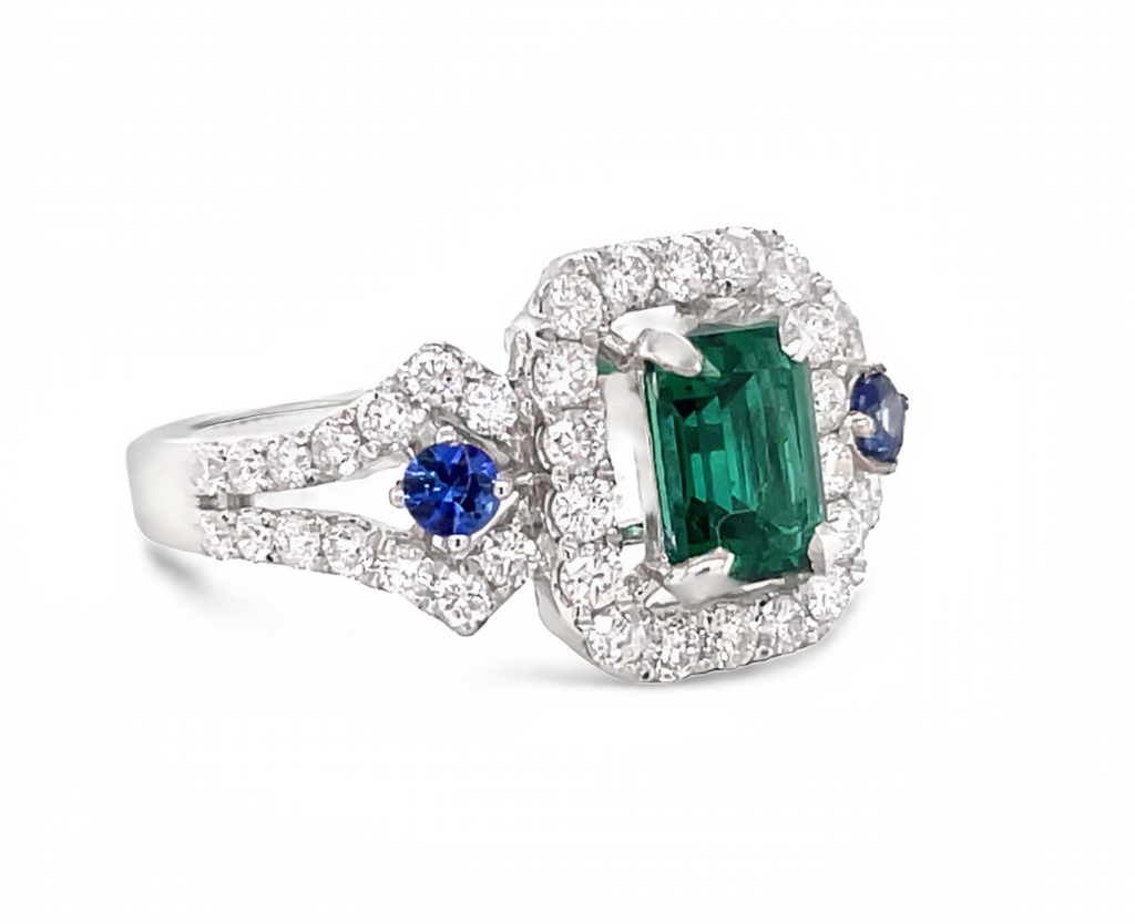 An emerald cut emerald 0.70 cts great hue color  Round 48 white diamonds 0.92 cts great quality  Two round blue sapphires  Set in 18k white gold  6.75 sizable ring   11.44 mm.