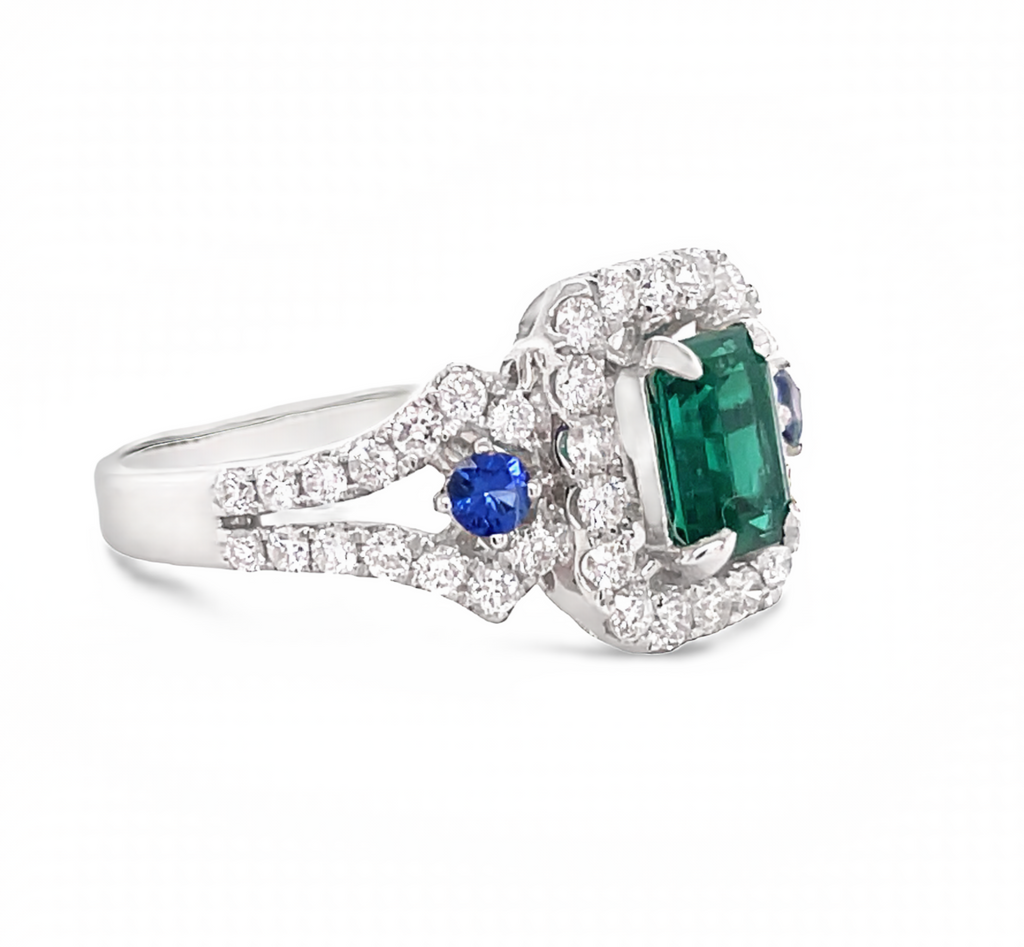 An emerald cut emerald 0.70 cts great hue color  Round 48 white diamonds 0.92 cts great quality  Two round blue sapphires  Set in 18k white gold  6.75 sizable ring   11.44 mm.