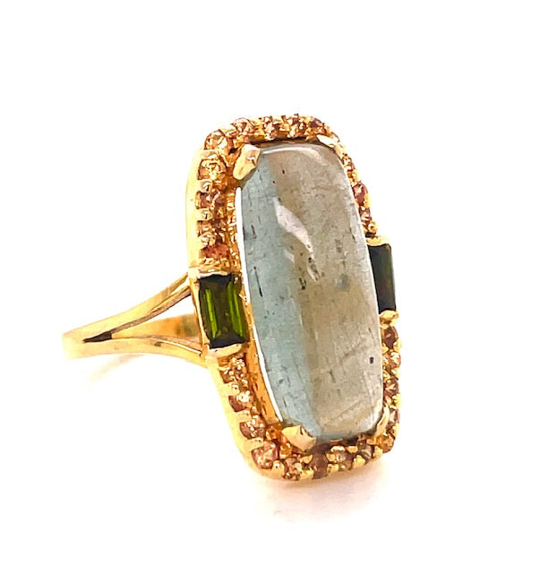 This timeless ring showcases a 20.00 x 12.00 mm majestic milky aquamarine cabochon surrounded by 0.02 cts twinkling yellow sapphires and 0.95 cts green tourmalines set in 14k yellow gold. The perfect combination of precious stones and handcrafted gold adds a luxe finish to any look.